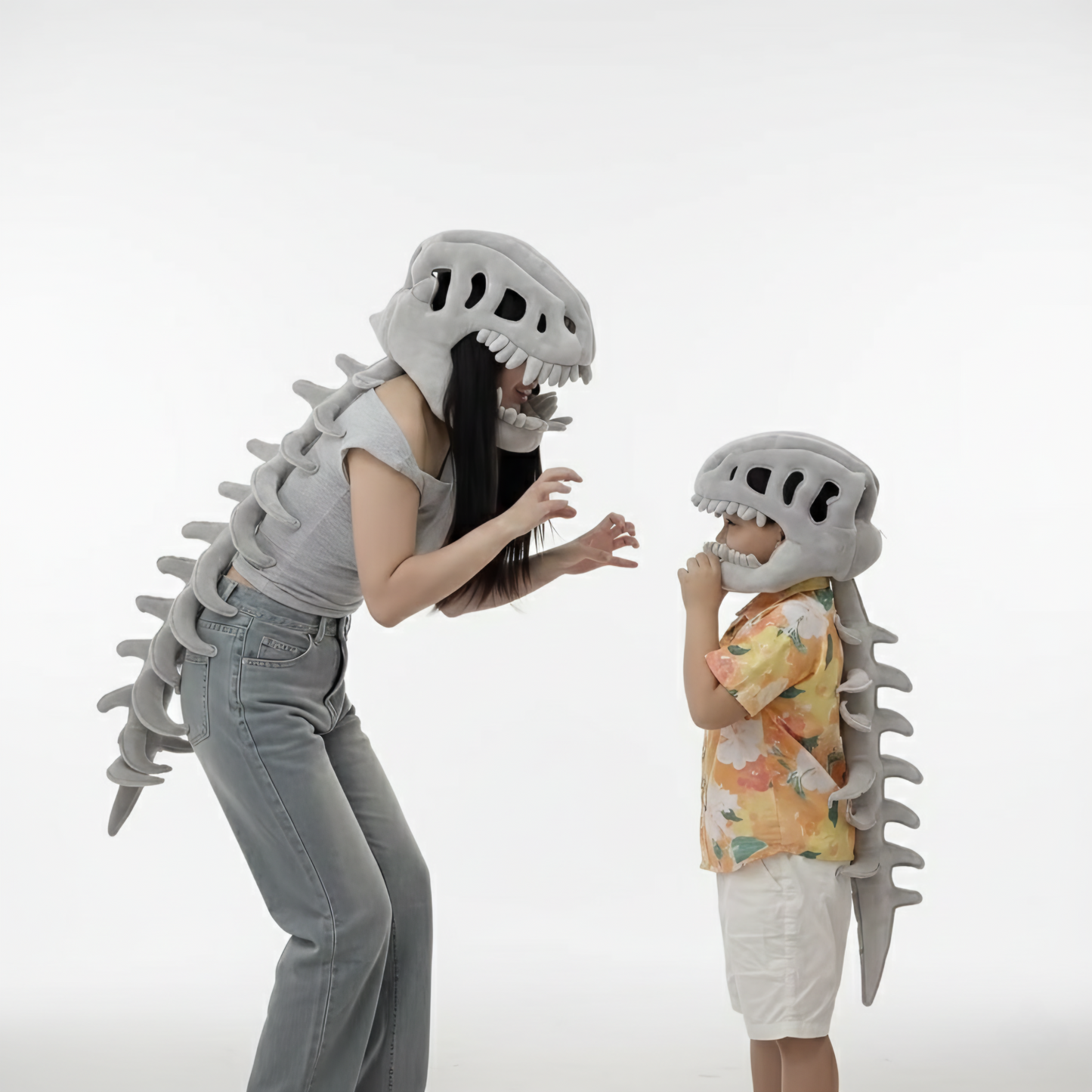 A woman and a young child are engaged in playful interaction in a bright white studio, both wearing matching plush grey dinosaur skeleton costumes. The dinosaur costume accessories feature a skull headpiece and a long, flexible spinal column down their backs. The woman leans forward, hands raised in a fun monster pose, while the child, wearing a yellow floral shirt and white shorts, stands facing her. This image is perfect for family bonding, parent-child costumes, Halloween apparel, fun dinosaur accessori
