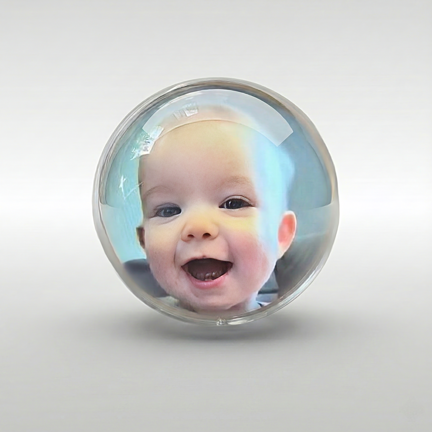 A high-resolution, centered studio image of a single clear glass or acrylic sphere floating slightly above a white surface. Inside the sphere, a projected or embedded image of a joyful, smiling baby's face is visible, appearing distorted slightly by the curved surface. The image is optimized for keywords such as holographic photo sphere, digital photo display globe, crystal ball photo frame, baby's first picture display, novelty photo gift, and modern memory display.