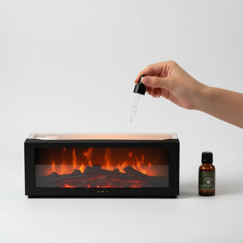 Hand holding a dropper over an electric fireplace with a bottle of oil next to it on a white background