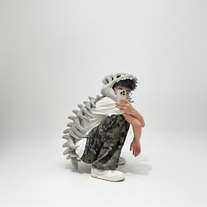 A young person is photographed in a studio against a plain white background, crouching low while wearing a plush grey dinosaur skeleton costume. The costume features a detailed skull hood and a long, articulated spinal column running down their back. The person is dressed in a white t-shirt, camouflage cargo pants, and white sneakers, and is holding a smartphone to take a selfie or mirror photo, with the phone obscuring part of their face. This image is suitable for costume design, casual fashion, unique a